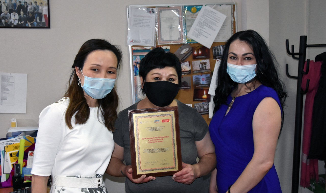 Head of NGO “Doverie plus” Roza Oleinikova with GHRCCA team. Three masked people holding a plaque/certificate