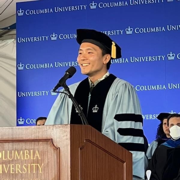 YG in a cap and gown at graduation, speaking at the podium