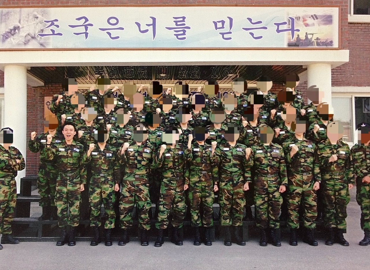 A group of South Korean Army trainees standing under a banner with Korean text that reads, ‘Your country believes in you’ Military photo with YG and other troops in South Korea