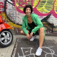 A woman with curly hair in a green blazer crouches in front of a wall painted with graffiti