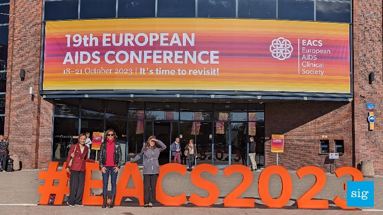 a building with a banner for the 19th european AIDS conference on it. three women are looking at the camera in front of the building