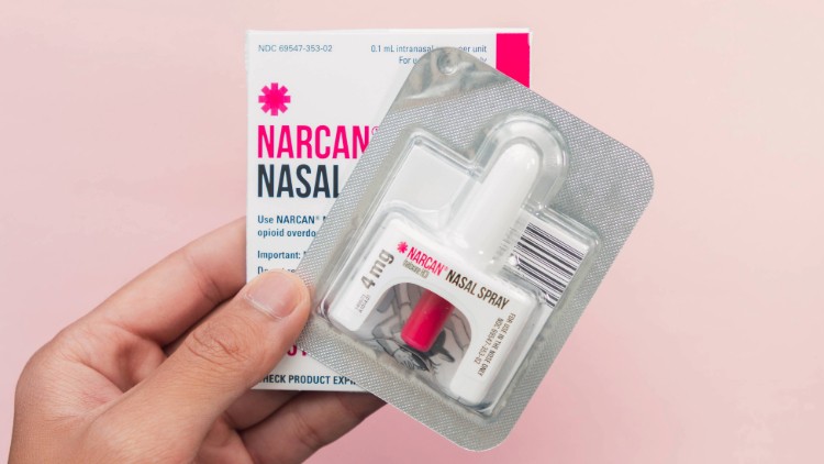 a package of Narcan nasal spray still in its foil packaging. Behind it the same hand holds up the box the medication was in
