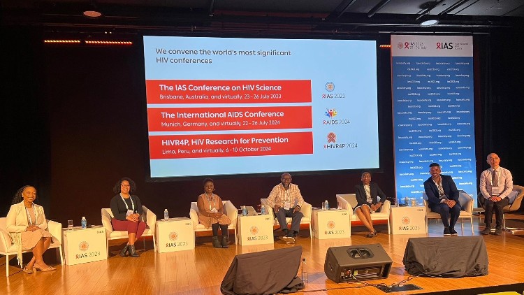 From left to right: Megan Threats, Jaih Craddock, Caroline Kingori, Peter Memiah, Melonie Walcott, Omar Martinez, Elwin Wu. Photo courtesy of Caroline Kingori. seven people sitting in chairs on a stage with a screen above them. The signs indicate they are at IAS 2023.