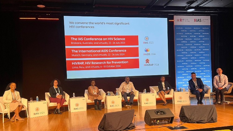 seven people sitting in chairs on a stage with a screen above them. The signs indicate they are at IAS 2023.