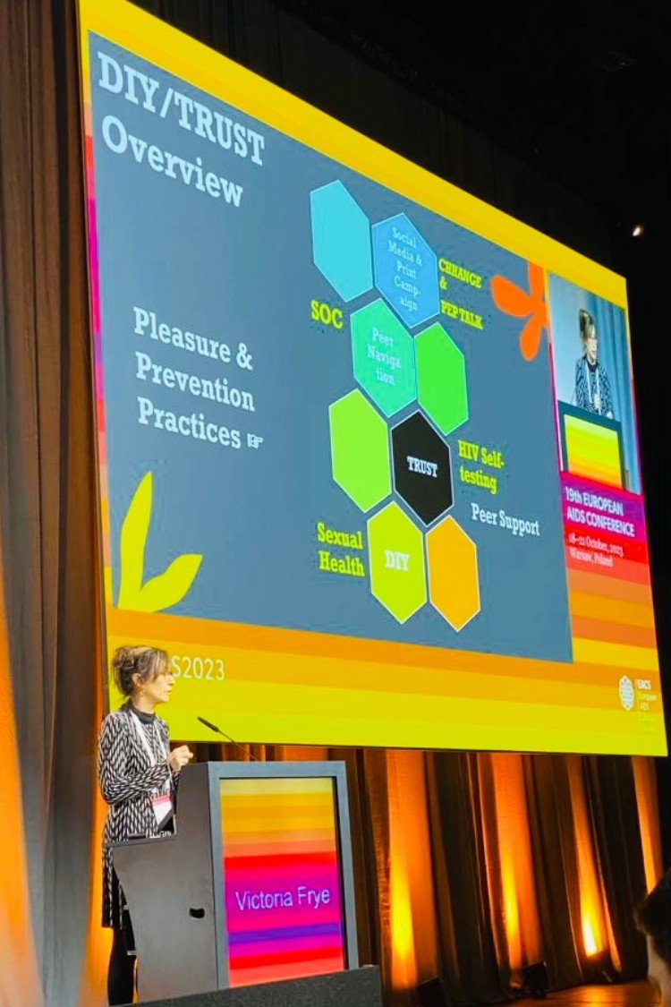a woman at a lectern in front of a screen that says "DIY/Trust Overview: Pleasure & Prevention Practices"