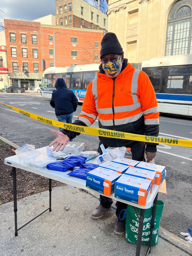 A man in an orange reflective jacket and a black hat stands behind a table of supplies on a city street