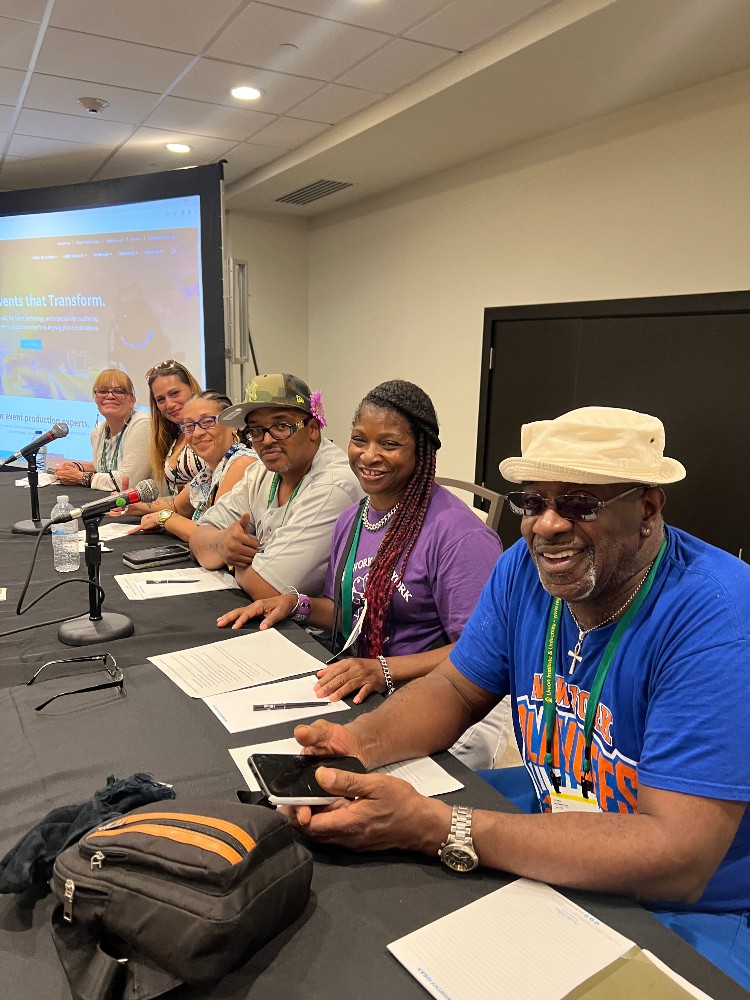 Will and the other peer researchers also presented their Labor of Love Project at the National Harm Reduction Conference in Puerto Rico. Pictured right to left from Will: Althea Matthews, Hiawatha Collins, Jeannette Mathews, Rachel Arana, and Marilyn Reyes. A group of people sitting at a conference table with microphones in front of them