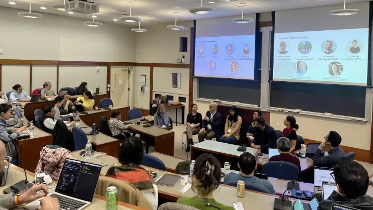 A large classroom of people taking notes on laptop computers. They're looking at a panel of six people on chairs at the front of the room, accompanied by a moderator.