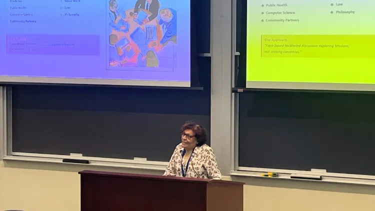 A woman in a patterned shirt stands at a lectern. Behind her are two large projection screens.