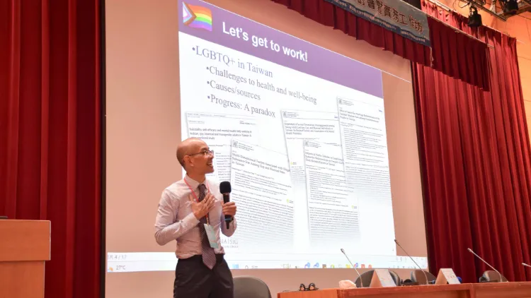A man in a tie stands in front of a projected slide holding a microphone