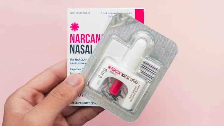 a package of Narcan nasal spray still in its foil packaging. Behind it the same hand holds up the box the medication was in