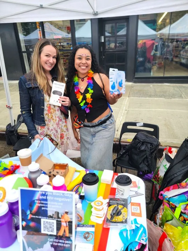 Two women smile at the camera from behind a table of colorful products. One of them is wearing a rainbow lei.