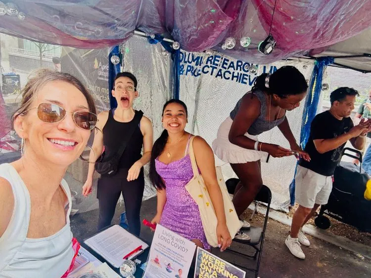 A wide selfie with the camera holder smiling and people in the background working and making faces at the camera