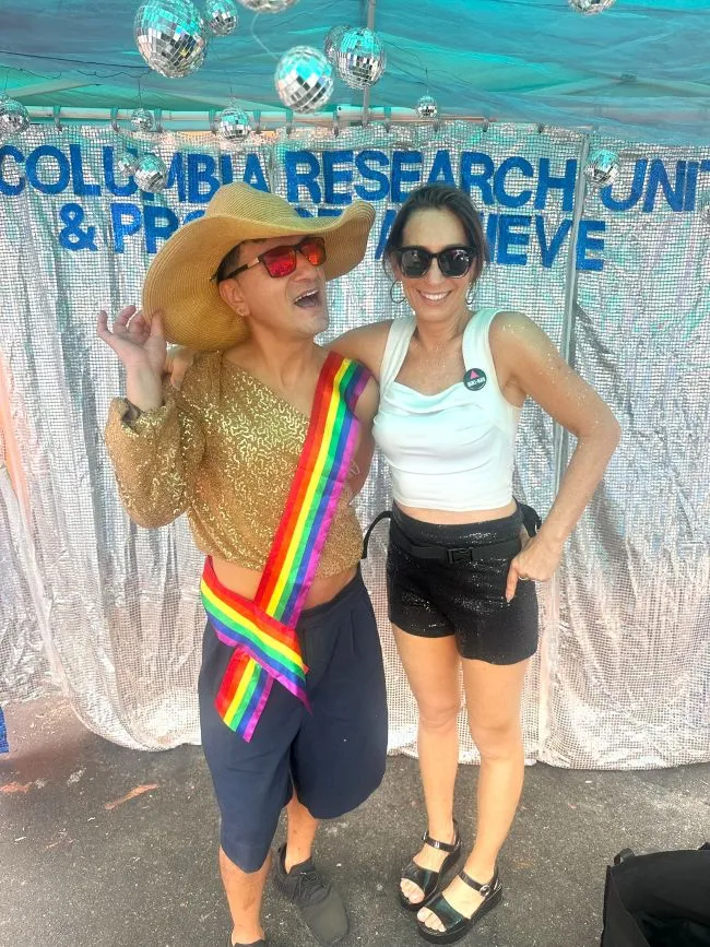 A man in a sparkly top wearing a rainbow sash poses with Victoria Frye