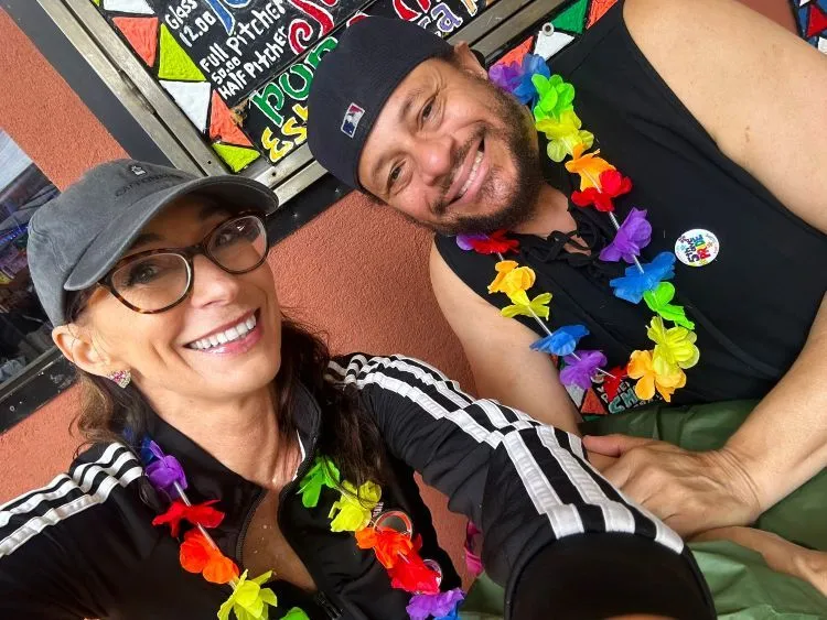 A man in a backward baseball cap and a woman in a baseball cap smile at the camera for a selfie. They are boht wearing rainbow leis.
