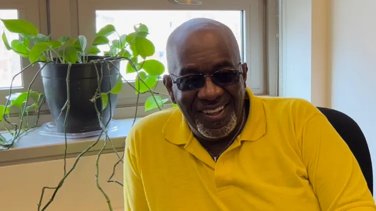 A man in a yellow shirt and glasses smiles. A window is behind him